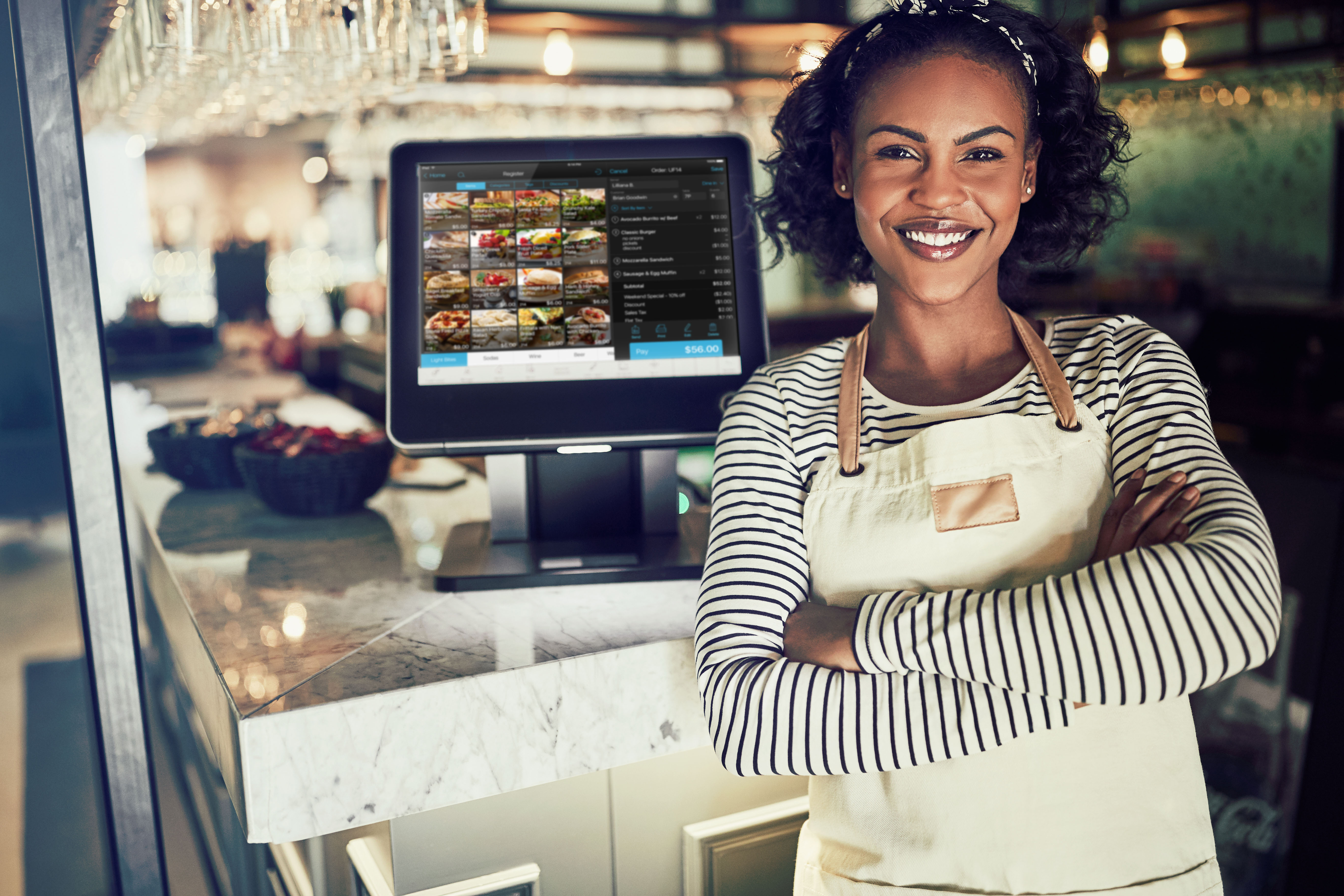 Young African waitress wearing an apron and smiling while standing by a point of sale terminal in a restaurant